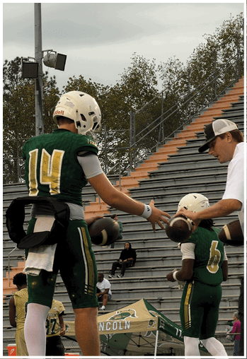 Several people wearing green and yellow football uniforms and helmets are raising their arms outdoors near a large tree. Description generated by AI
