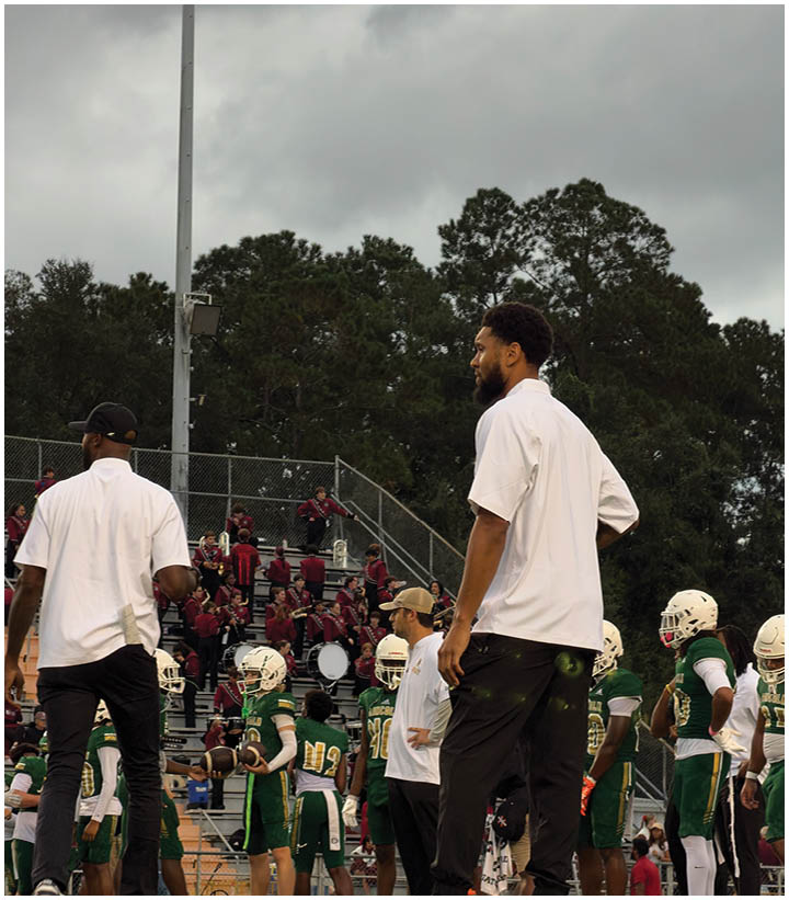 Two people in white shirts stand on a sports field near a tall light pole with a group of people seated in bleachers behind them under a cloudy sky. Description generated by AI