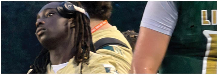 A person wearing headphones and a light-colored jacket is looking to the side while standing near another person in a green sports jersey with the word Lincoln and the number 13 on the back. Description generated by AI