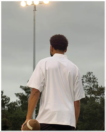 A person with short curly hair wearing a white shirt is holding a football and looking up at bright stadium lights against a cloudy sky with trees in the background. Description generated by AI