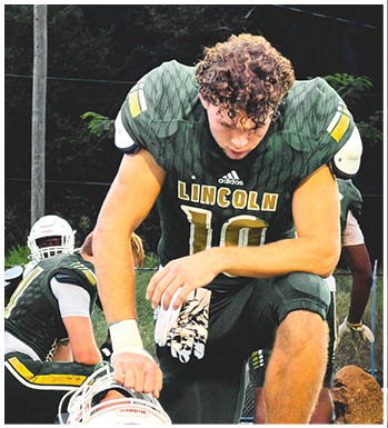 A person in a green football uniform with “LINCOLN" on the chest is kneeling on a field near a goalpost while another person in similar attire is crouched behind them. Description generated by AI