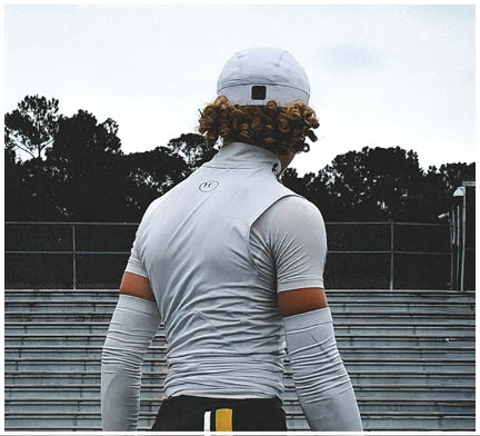 A person wearing a white athletic outfit and cap stands in front of empty bleachers with trees and a light pole in the background. Description generated by AI
