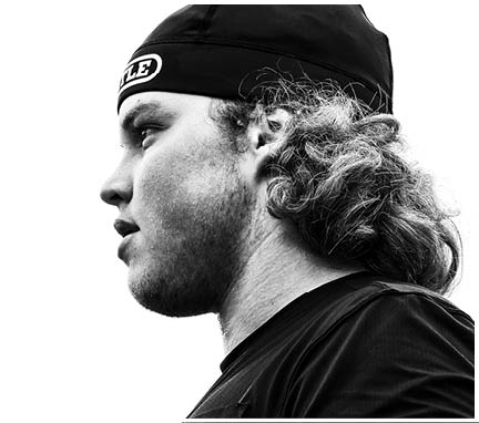 Person with long curly hair wearing a backward cap and a dark shirt looking to the left in a high-contrast black and white photo. Description generated by AI
