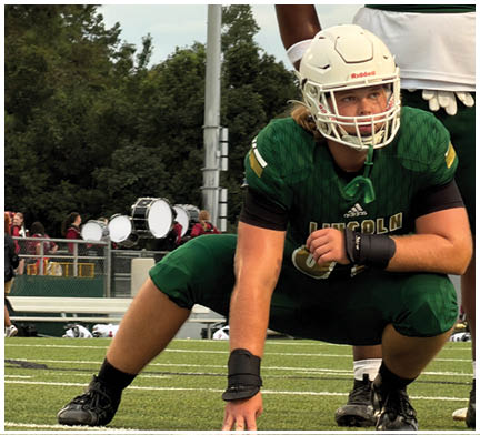Two people wearing green football uniforms and white helmets are outdoors with trees and a cloudy sky in the background. Description generated by AI