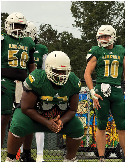 Three people wearing green football uniforms with white helmets stand and sit near a chain-link fence with trees and a cloudy sky in the background. Description generated by AI