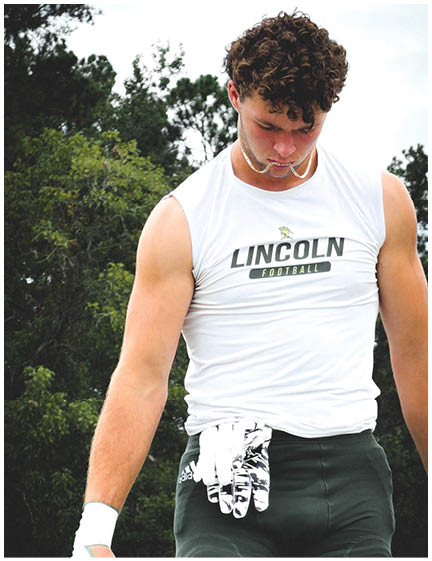An adult with curly hair wearing a sleeveless white shirt with “Lincoln Football" printed on it is looking down while standing outdoors with trees in the background. Description generated by AI