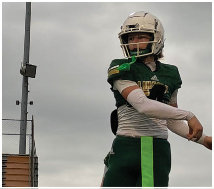 A person wearing a white football helmet and green jersey is looking to the side under a cloudy sky near a tall light pole. Description generated by AI