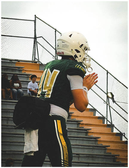 A person wearing a football uniform and helmet stands on the sidelines of a stadium with empty bleachers in the background. Description generated by AI