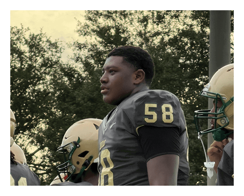 A person wearing a dark sports jersey with the number 58 stands outdoors near others in helmets, with trees and a cloudy sky in the background. Description generated by AI