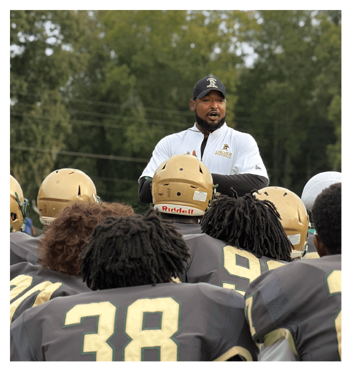 A person wearing a white sports jacket and a black cap is speaking to a group of people wearing gold football helmets outdoors with trees in the background. Description generated by AI