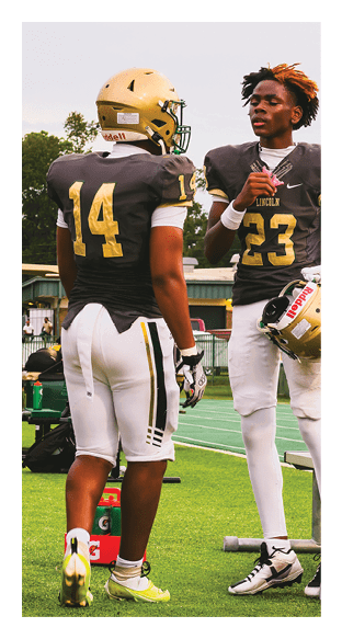 Two people wearing dark football uniforms and yellow helmets are standing outdoors, with one person’s jersey number 14 visible. Description generated by AI