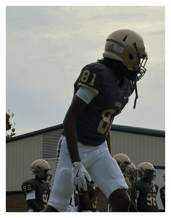 A person wearing a football uniform with the number 81 and a helmet is standing outdoors near other players in similar gear with a building in the background. Description generated by AI