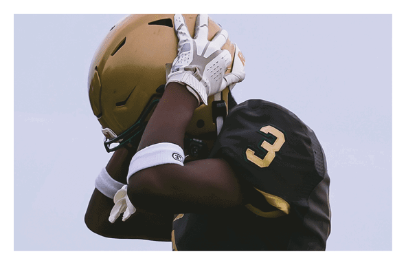 A person wearing a black and gold football uniform with the number three is adjusting their helmet against a plain sky background. Description generated by AI