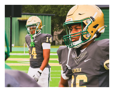 Two people wearing football helmets and dark jerseys with gold helmets stand on a football field with green turf and a green building in the background. Description generated by AI