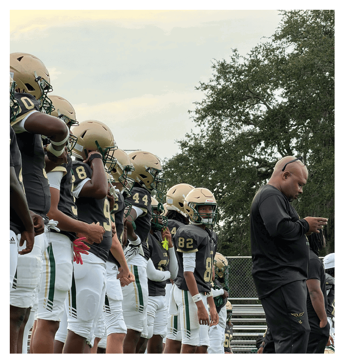 A group of football players in helmets and uniforms stand in a line on the field while a coach or staff member in black athletic wear looks at a phone nearby. Description generated by AI
