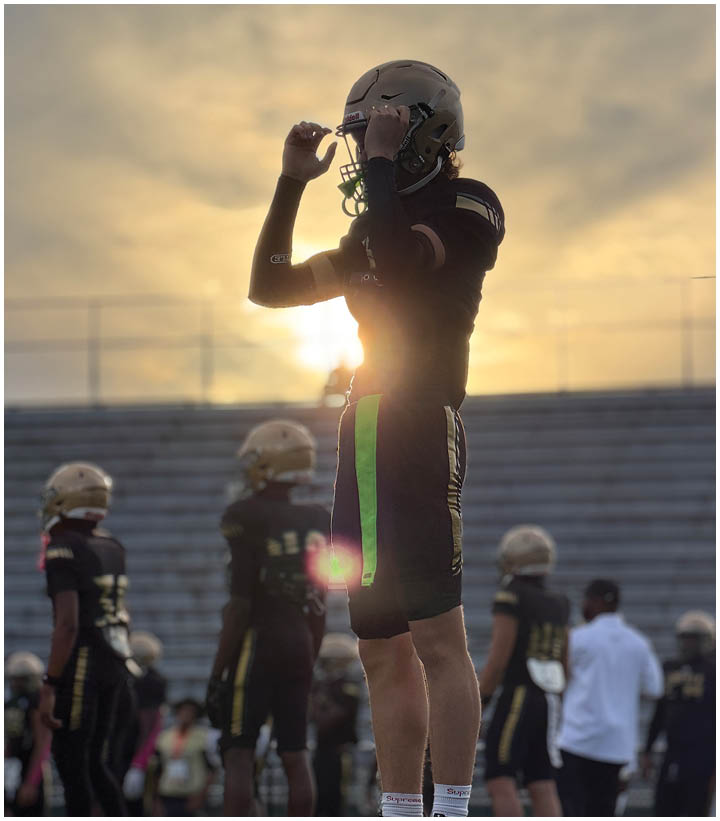 Adults in football uniforms stand on a field with stadium bleachers in the background during sunset. Description generated by AI