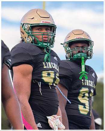 Two people wearing black football uniforms with gold helmets and pink arm tape stand side by side on a field. Description generated by AI