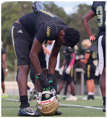 A person wearing sports gear is bending down on a football field, holding a gold football helmet with the brand name Riddell visible. Description generated by AI