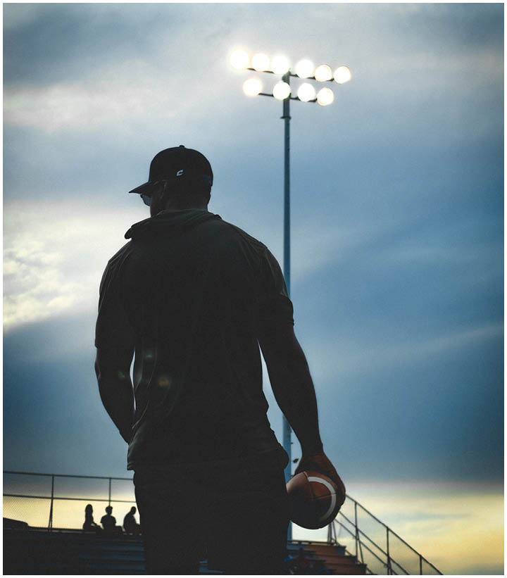 A person wearing a cap and dark clothing is holding a football while standing near bleachers under a cloudy sky at sunset. Description generated by AI
