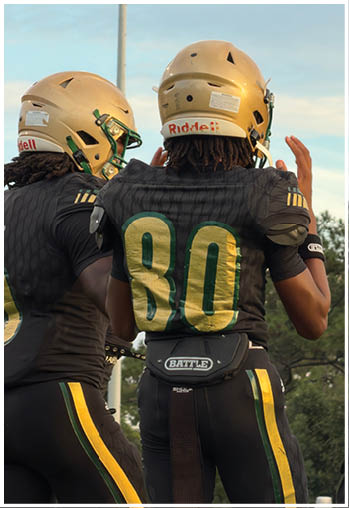 Two people wearing black sports uniforms with yellow and green stripes are standing outdoors, one with a football belt and towel attached to their waist. Description generated by AI
