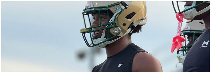 A person wearing a black athletic outfit with yellow stripes stands in the foreground while another person in a helmet is blurred in the background on a sports field. Description generated by AI