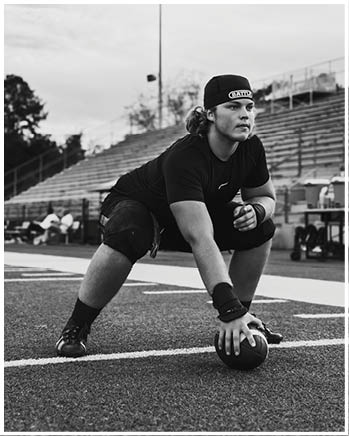 A person wearing athletic clothing and a headband is crouched on a football field holding a football in a ready position with empty bleachers in the background. Description generated by AI