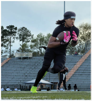 An adult wearing black athletic clothing and bright yellow cleats is running on a football field while holding a football and wearing pink gloves. Description generated by AI
