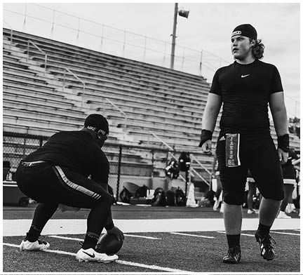 Two people on a football field are preparing for a play with one person holding the football on the ground and the other standing nearby wearing a towel with the number 61. Description generated by AI