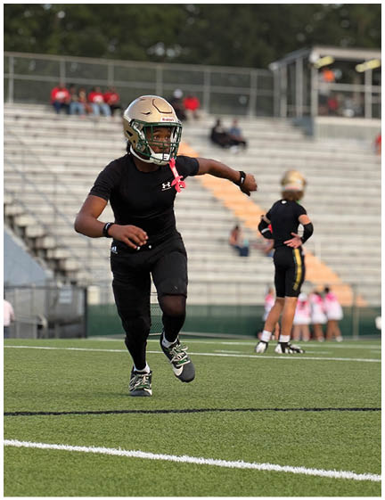 A person wearing a gold helmet and black athletic clothing is running on a football field with other people and empty bleachers in the background. Description generated by AI