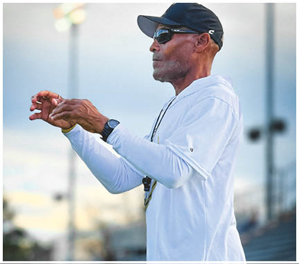 A senior person wearing a white long-sleeve shirt and dark pants is standing outdoors, gesturing with their hands as if giving instructions or coaching. Description generated by AI