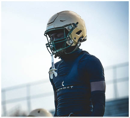 Two people wearing football uniforms stand on a field with bleachers in the background, one in a dark outfit with a glove hanging from the waist and the other wearing a helmet. Description generated by AI