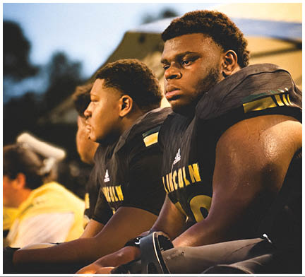 Three people wearing dark sports uniforms with shoulder pads sit closely together, looking intently in the same direction, with a blurred outdoor background. Description generated by AI