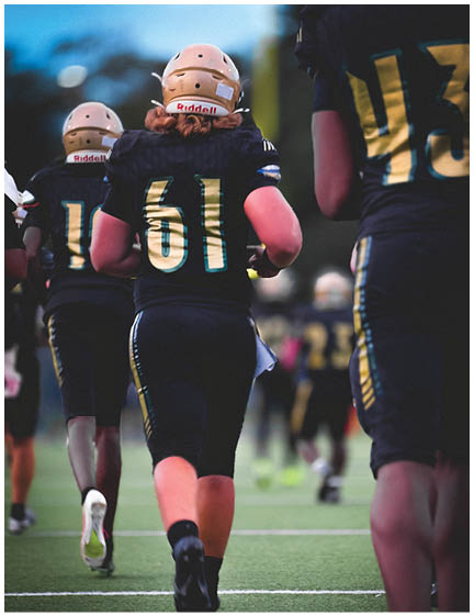 People wearing football uniforms with helmets are walking on a field, with one person in the center wearing number 61. Description generated by AI