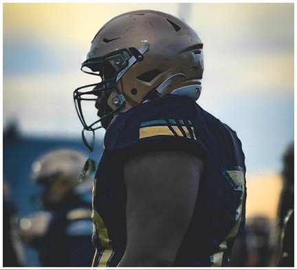 A person wearing a football helmet and dark uniform is standing on a field during sunset with blurred figures in the background. Description generated by AI