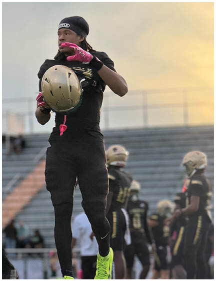 A person in black athletic gear and bright yellow shoes is holding a gold football helmet on a field with other similarly dressed people and empty bleachers in the background. Description generated by AI