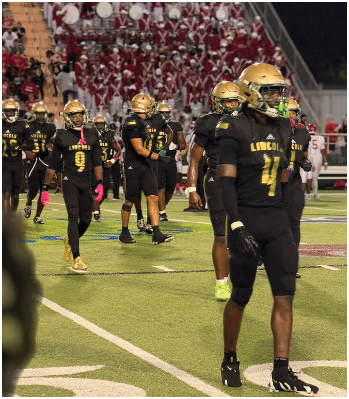 Several people in black and gold football uniforms stand on a field with a marching band in red uniforms visible in the background. Description generated by AI