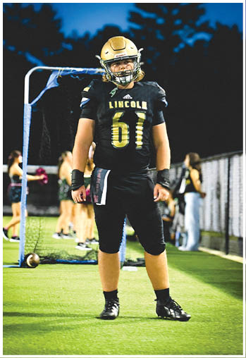 A person wearing a black football uniform with the number 61 and the word Lincoln stands on a green field with a football on the ground nearby and blurred people in the background. Description generated by AI