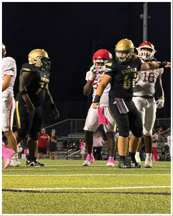 People wearing football uniforms stand on a field with artificial turf under stadium lights. Description generated by AI