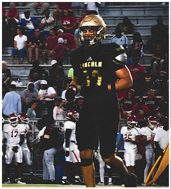 A person in a black football uniform with the word Lincoln and the number 13 is on a football field while a group of people in white uniforms and helmets stand on the sidelines. Description generated by AI
