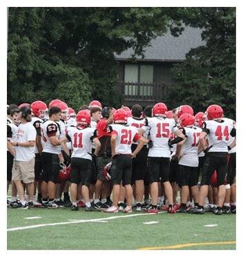 A group of people wearing football uniforms and helmets are gathered closely together on a sports field, appearing to be in a team huddle or discussion. Description generated by AI