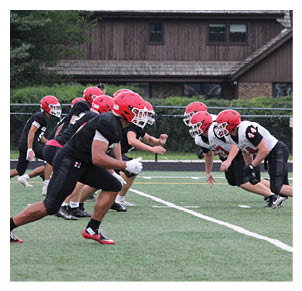 Two opposing football teams wearing red helmets and black and white uniforms are lined up facing each other on a green field preparing for a play. Description generated by AI