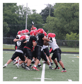 A group of people wearing red helmets and black or white sports uniforms are engaged in a football play on a green field with trees and a fence in the background. Description generated by AI