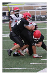 Two people wearing red helmets and black sports uniforms are tackling another person on a football field near the sideline. Description generated by AI