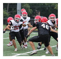 People wearing helmets and football uniforms are actively playing on a grassy field with one person running with the ball while others attempt to block or tackle. Description generated by AI