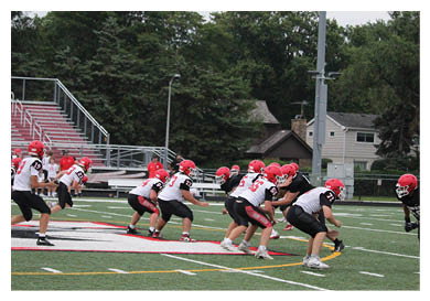 Players in red helmets and black and white uniforms are lined up on a football field preparing for a play during a game. Description generated by AI