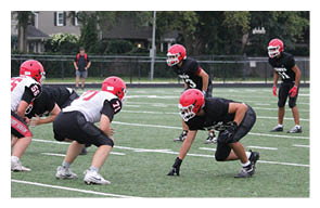 Two people wearing black sports uniforms and red helmets are positioned on a football field with trees and a fence in the background. Description generated by AI