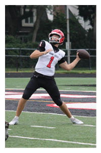A person wearing a red helmet and white jersey with the number one is preparing to throw a football while other players and a referee stand nearby on a football field. Description generated by AI