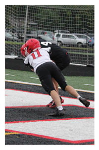Two people in football gear are engaged in a tackle near the end zone on a football field with a goalpost and parked cars in the background. Description generated by AI
