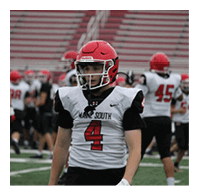 A person wearing a red helmet and white football jersey with the number 4 stands on a football field with other players and a referee in the background. Description generated by AI
