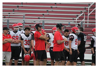 People wearing sports uniforms and red shirts stand on the sidelines of a football field holding helmets. Description generated by AI